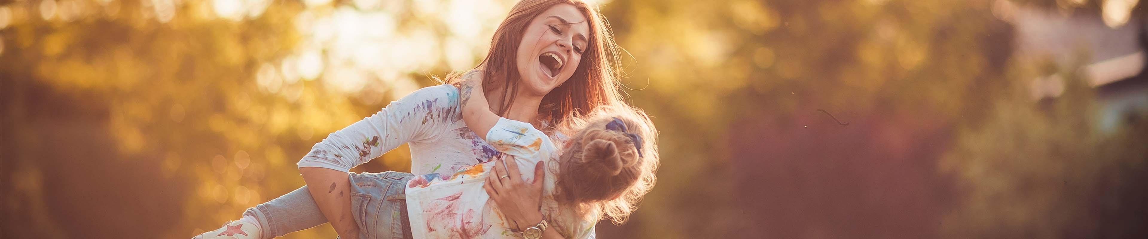 A white woman covered in paint happily swings a white little girl also covered in paint around their yard under the setting sun.