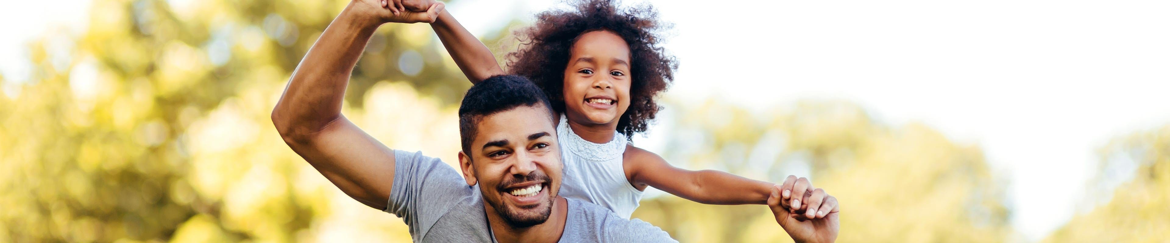 Black father with short-cropped hair carrying his black daughter on his back playing airplane together.